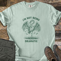 I'm Not Being Dramatic Comfort Colors T-shirt in Bay, laid flat. A green etched flamingo stands on one leg in rippling water, wings spread and feathers falling, between the words I'm Not Being Dramatic. Olive cargo pants and brown hiking boots sit on a rustic wood floor.