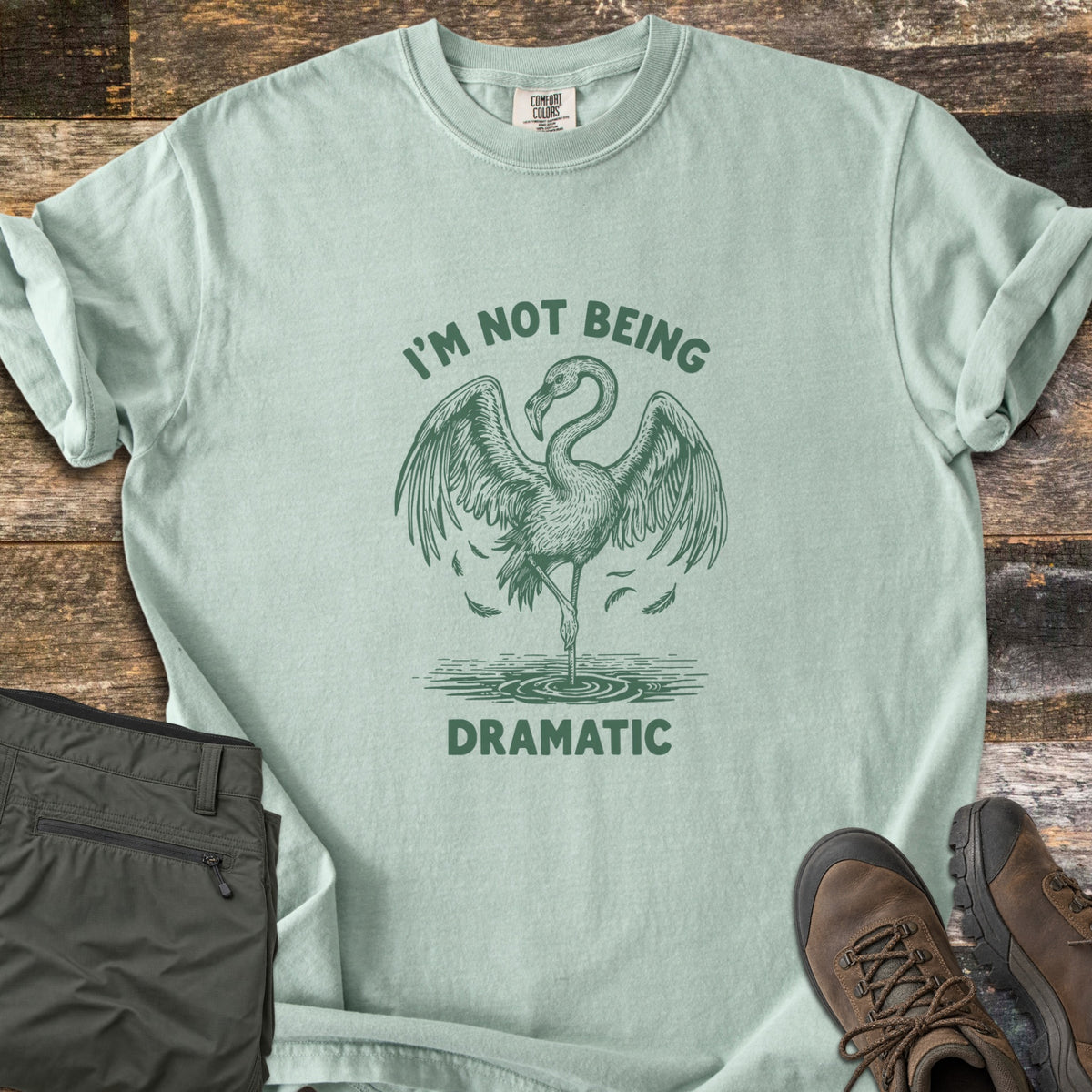 I'm Not Being Dramatic Comfort Colors T-shirt in Bay, laid flat. A green etched flamingo stands on one leg in rippling water, wings spread and feathers falling, between the words I'm Not Being Dramatic. Olive cargo pants and brown hiking boots sit on a rustic wood floor.