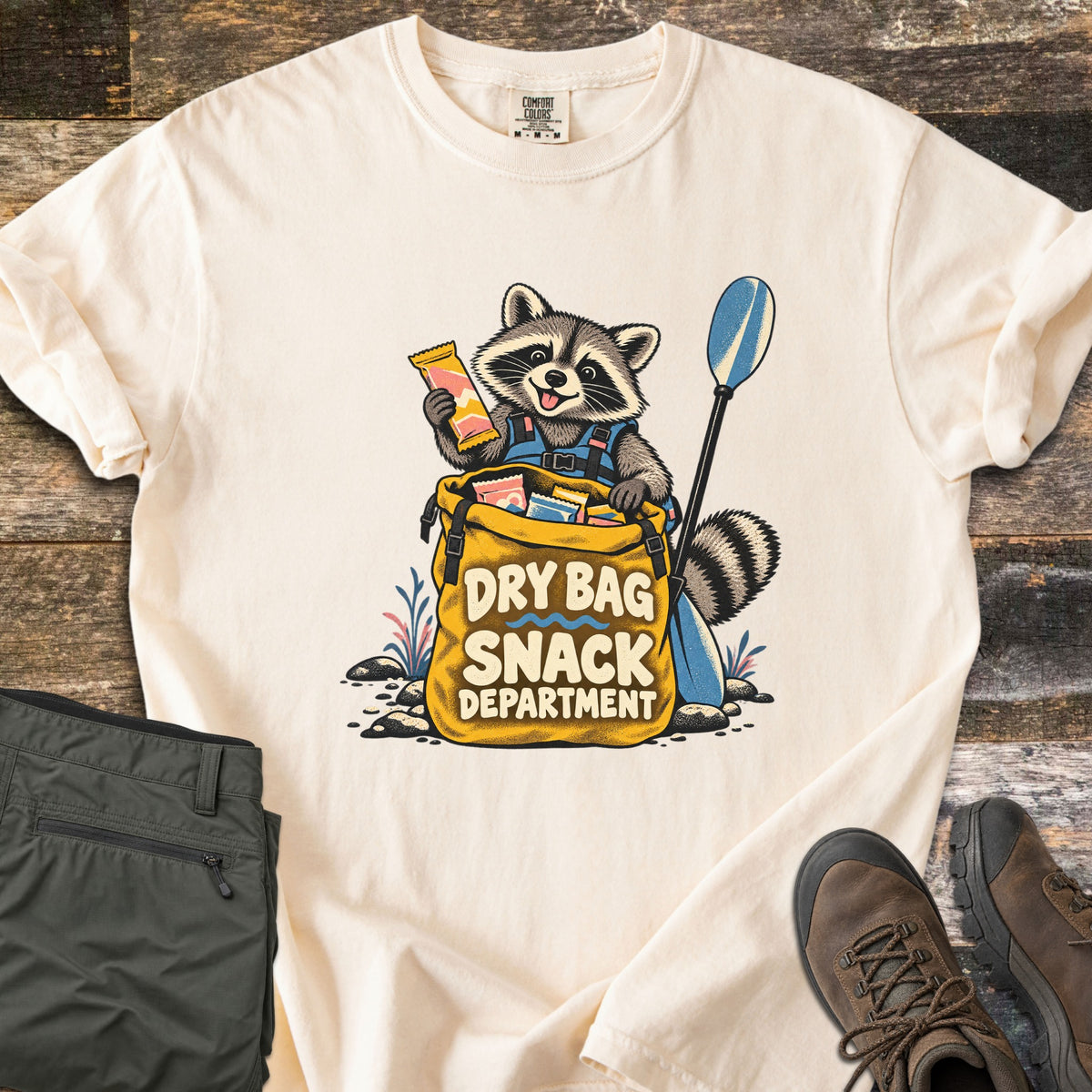 Snack Department Comfort Colors T-shirt in Ivory, laid flat. A raccoon in a blue life vest holds a snack bar over a yellow dry bag full of snacks, a paddle beside it, with the words Dry Bag Snack Department. Olive pants and brown hiking boots sit on wood planks.