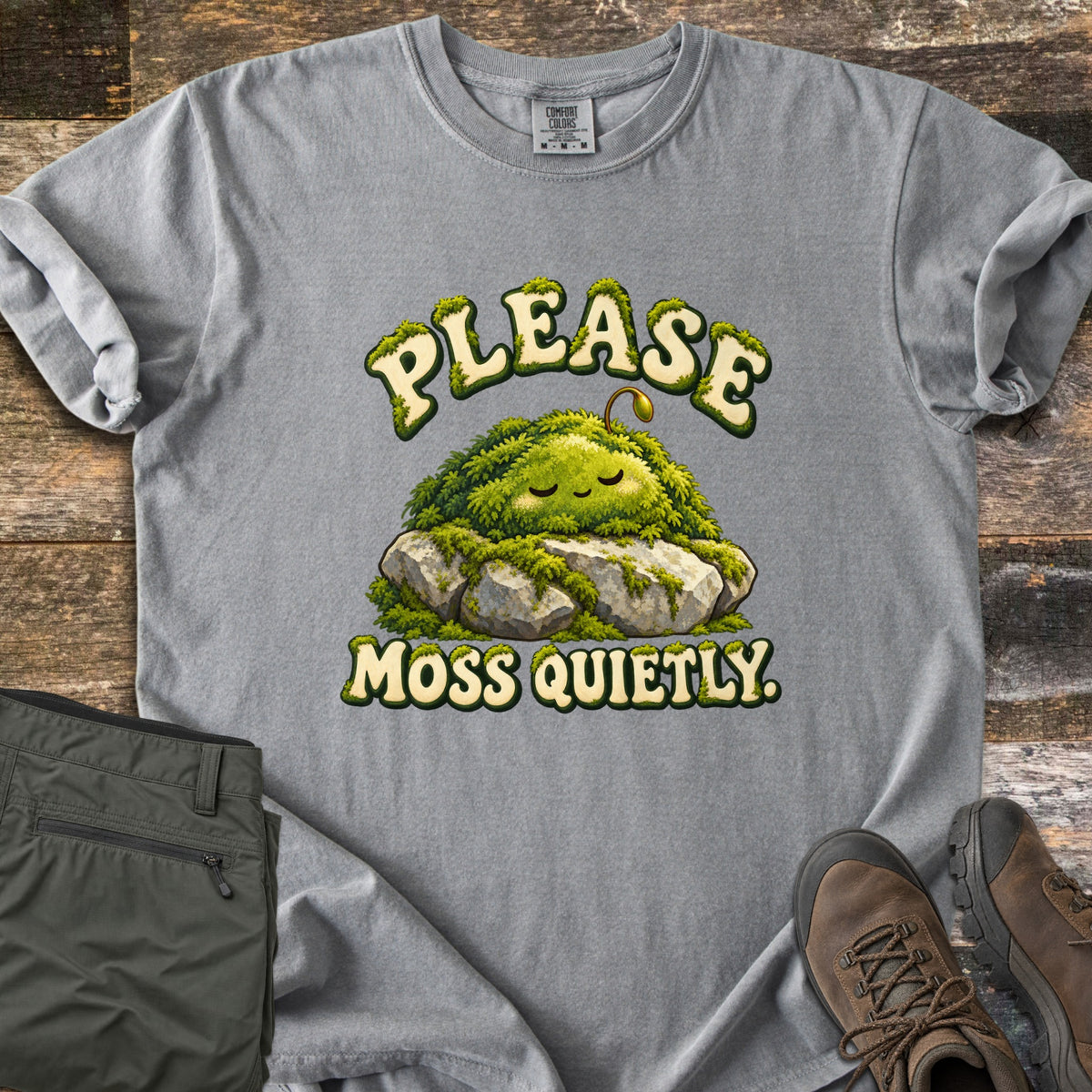 Please Moss Quietly Comfort Colors T-shirt in Grey, laid flat. A sleepy, smiling clump of moss with a tiny sprout rests on gray rocks, with PLEASE arched above and MOSS QUIETLY. below in moss-covered letters. Olive hiking pants and brown hiking boots lie on weathered wood.