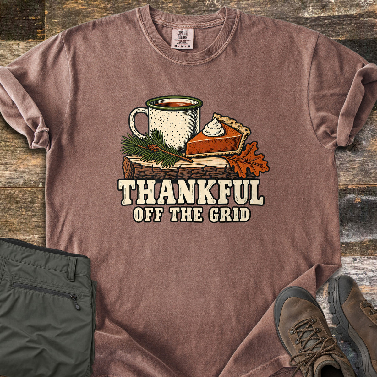 Thankful Off the Grid Comfort Colors T-shirt in Espresso, laid flat. A speckled enamel camp mug and a slice of pumpkin pie with whipped cream sit on a log with a pine sprig and an oak leaf above the words Thankful Off the Grid. Olive hiking pants and brown boots lie on weathered wood.