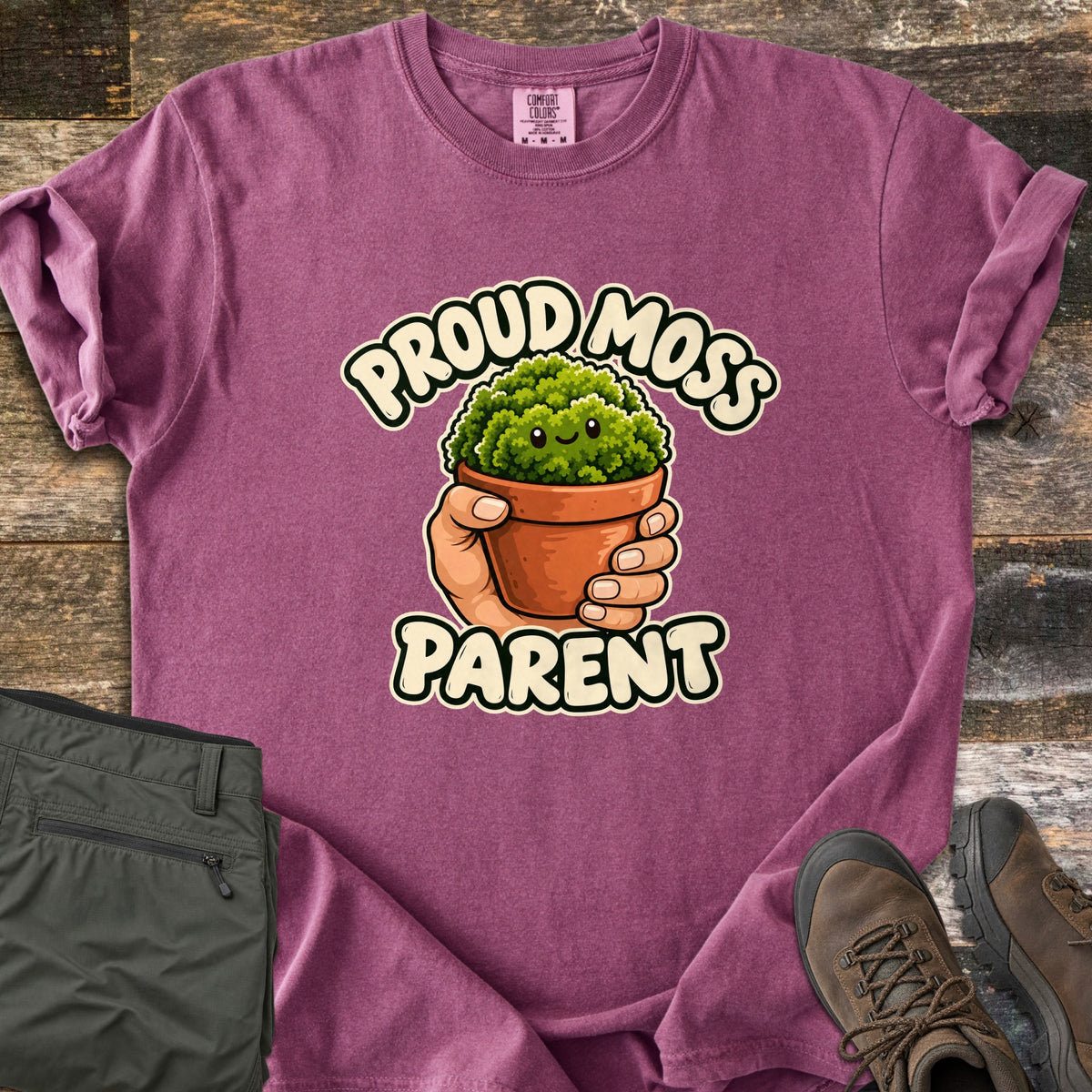 Proud Moss Parent Comfort Colors T-shirt in Berry, laid flat. A cartoon hand holds a terracotta pot of smiling green moss between the words Proud Moss Parent. Sleeves are rolled, with green hiking pants and brown hiking boots on a weathered wood background.