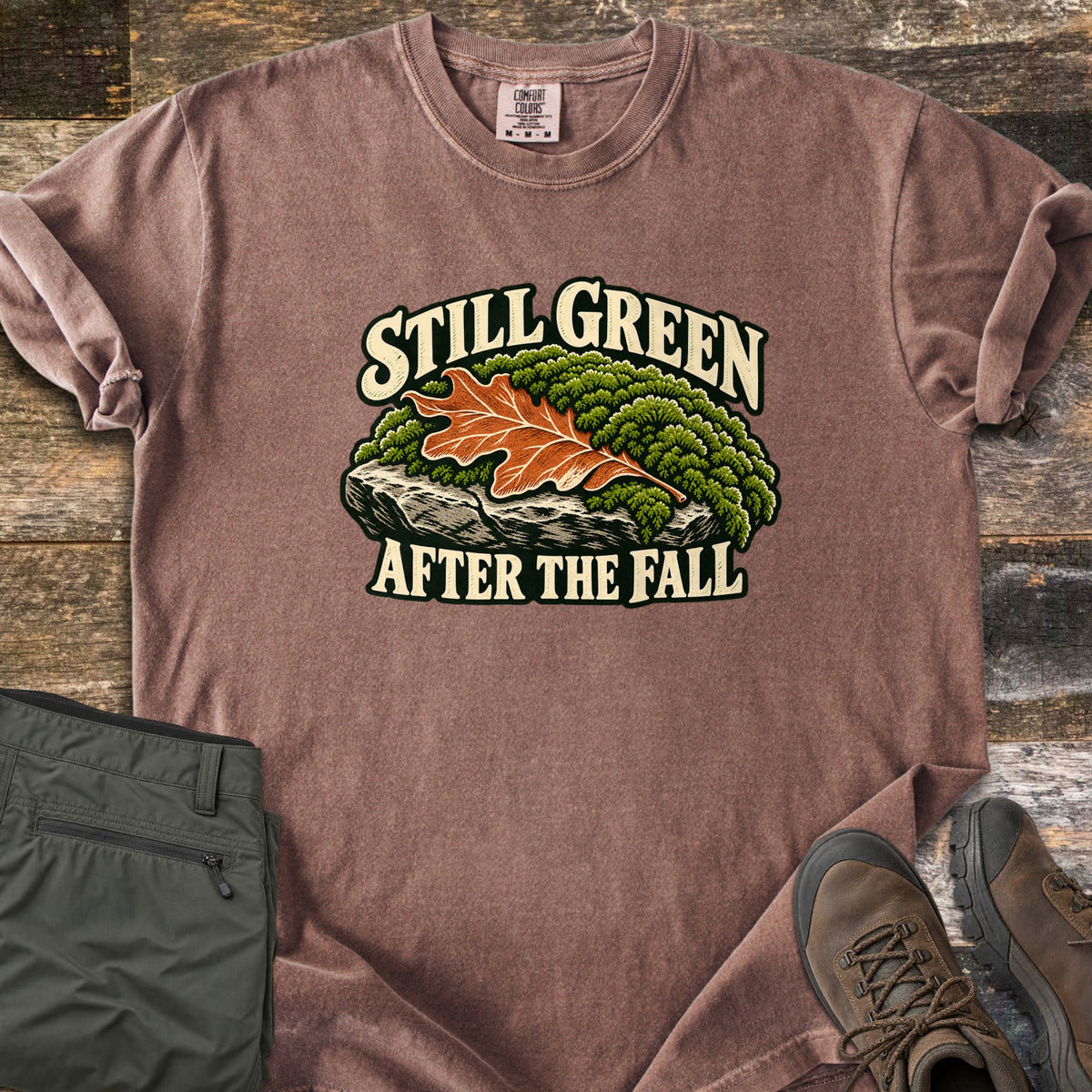 Still Green After the Fall Comfort Colors T-shirt in Espresso, laid flat. An orange oak leaf lies on green moss over a gray rock, with Still Green above and After the Fall below in cream letters. Folded olive hiking pants and brown hiking boots sit beside it on rustic wood planks.
