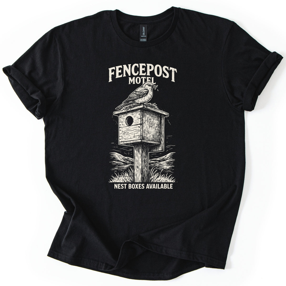 Fencepost Motel Comfort Colors T-shirt in Black, laid flat. A cream sketch of a bird perched on a wooden birdhouse atop a post amid grass and hills, with Fencepost Motel above and Nest Boxes Available below. Sleeves rolled, on a white background.