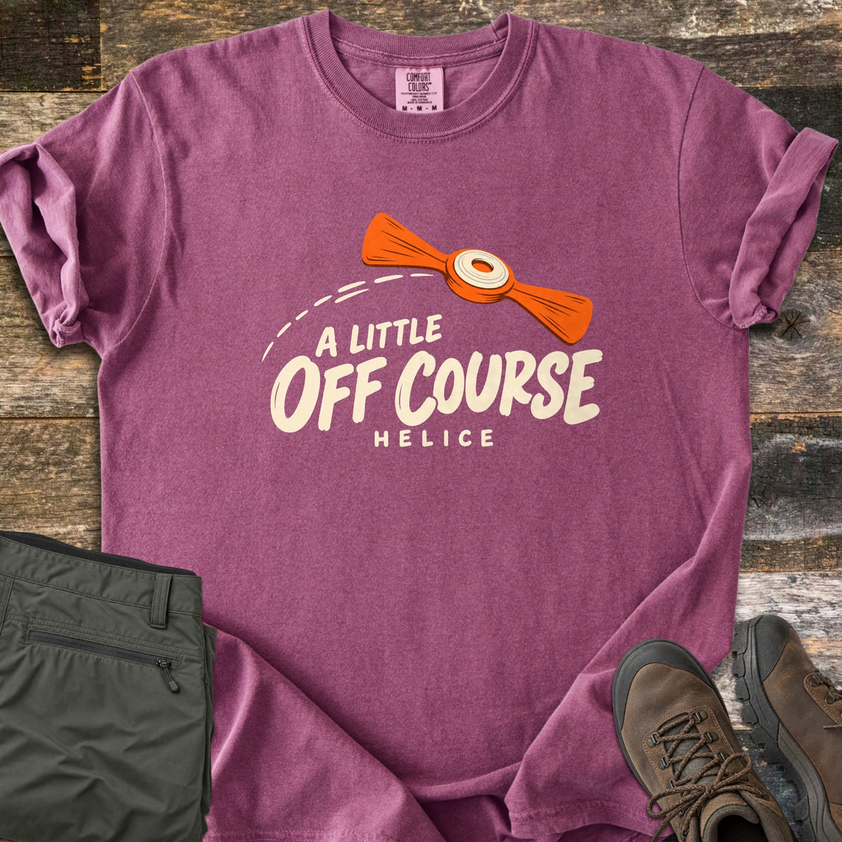 A Little Off Course Comfort Colors T-shirt in Berry, laid flat. An orange helice target with a white center flies along a dashed arc above cream text reading A Little Off Course and Helice. Folded olive hiking pants and brown hiking boots sit beside it on weathered wood.