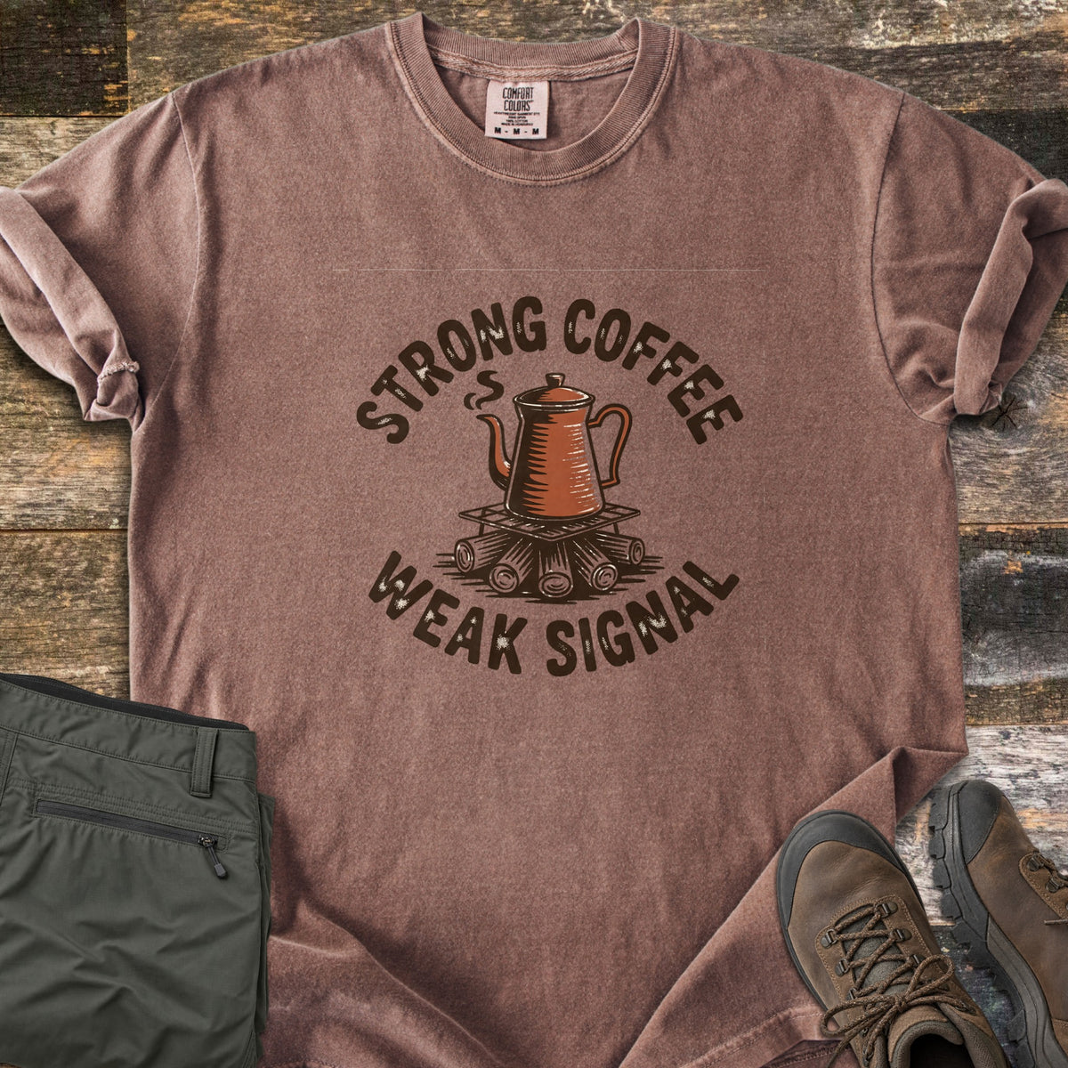 Strong Coffee Weak Signal Comfort Colors T-shirt in Espresso, laid flat. A steaming copper coffee pot sits on a grate over campfire logs, arched by the words Strong Coffee above and Weak Signal below. Folded olive hiking pants and brown hiking boots lie on weathered wood planks.