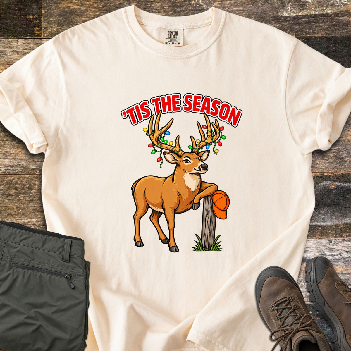 Buck Tis The Season Comfort Colors T-shirt in Ivory, laid flat. A cartoon buck with colorful Christmas lights wrapped in its antlers leans on a wooden post holding an orange cap, under red words 'Tis The Season. Olive pants and brown hiking boots lie on weathered wood.