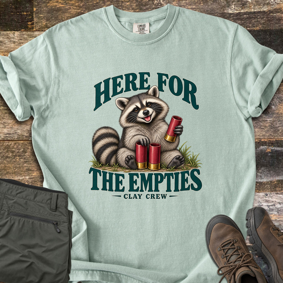 Here for the Empties Comfort Colors T-shirt in Bay, laid flat. A smiling raccoon sits in grass holding red shotgun shells between the words Here for the Empties, with Clay Crew below. Olive cargo pants and brown hiking boots lie beside it on weathered wood.
