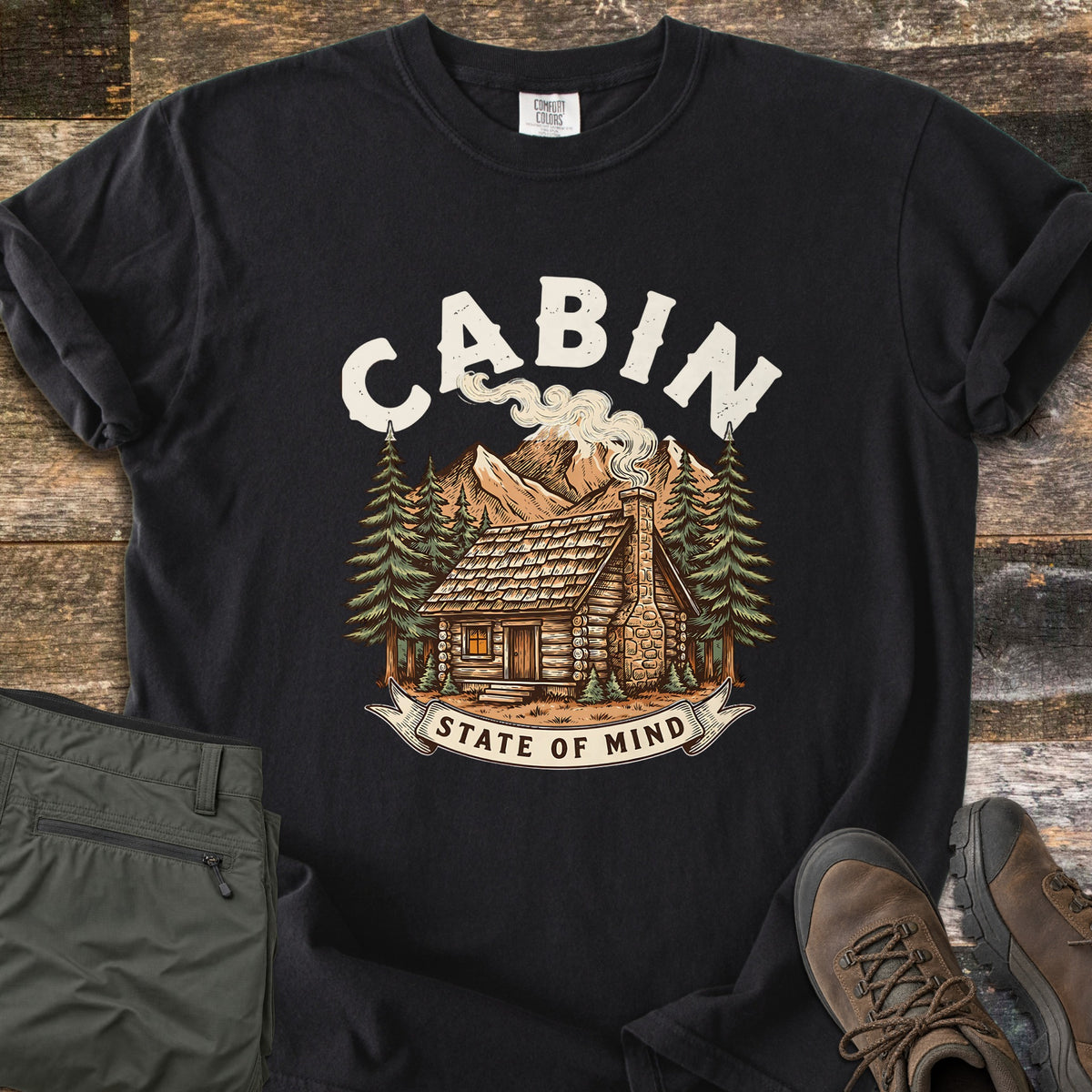 Cabin State Of Mind Comfort Colors T-shirt in Black, laid flat. A log cabin with a smoking stone chimney sits among pine trees and mountains under the arched word CABIN in cream, with a banner reading State Of Mind. Green pants and brown hiking boots lie beside it on weathered wood.