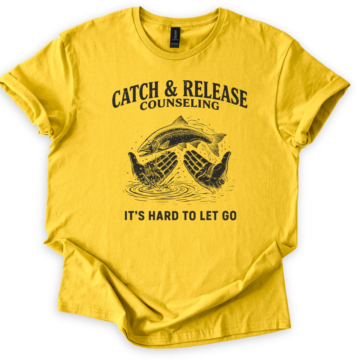 Catch & Release Counseling Comfort Colors T-shirt in Daisy, laid flat. Black print reads CATCH & RELEASE COUNSELING above an etched trout leaping from two open hands over rippling water, with IT'S HARD TO LET GO below. Sleeves are cuffed on a white background.