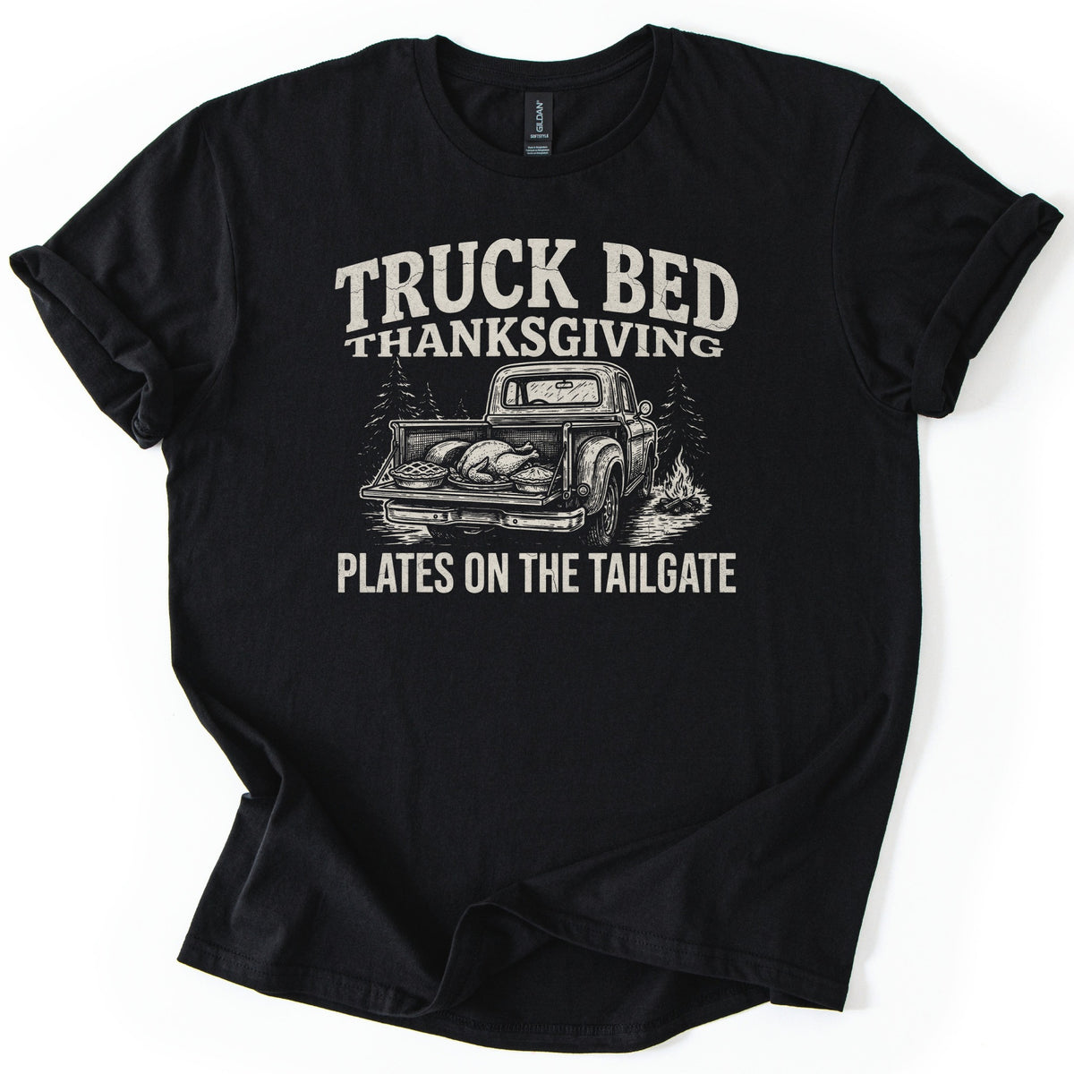 Truck Bed Thanksgiving Comfort Colors T-shirt in Black, laid flat. A cream print reads TRUCK BED THANKSGIVING over a vintage pickup with a roast turkey and a pie on its open tailgate beside pine trees and a campfire, above PLATES ON THE TAILGATE. Sleeves cuffed, on white.
