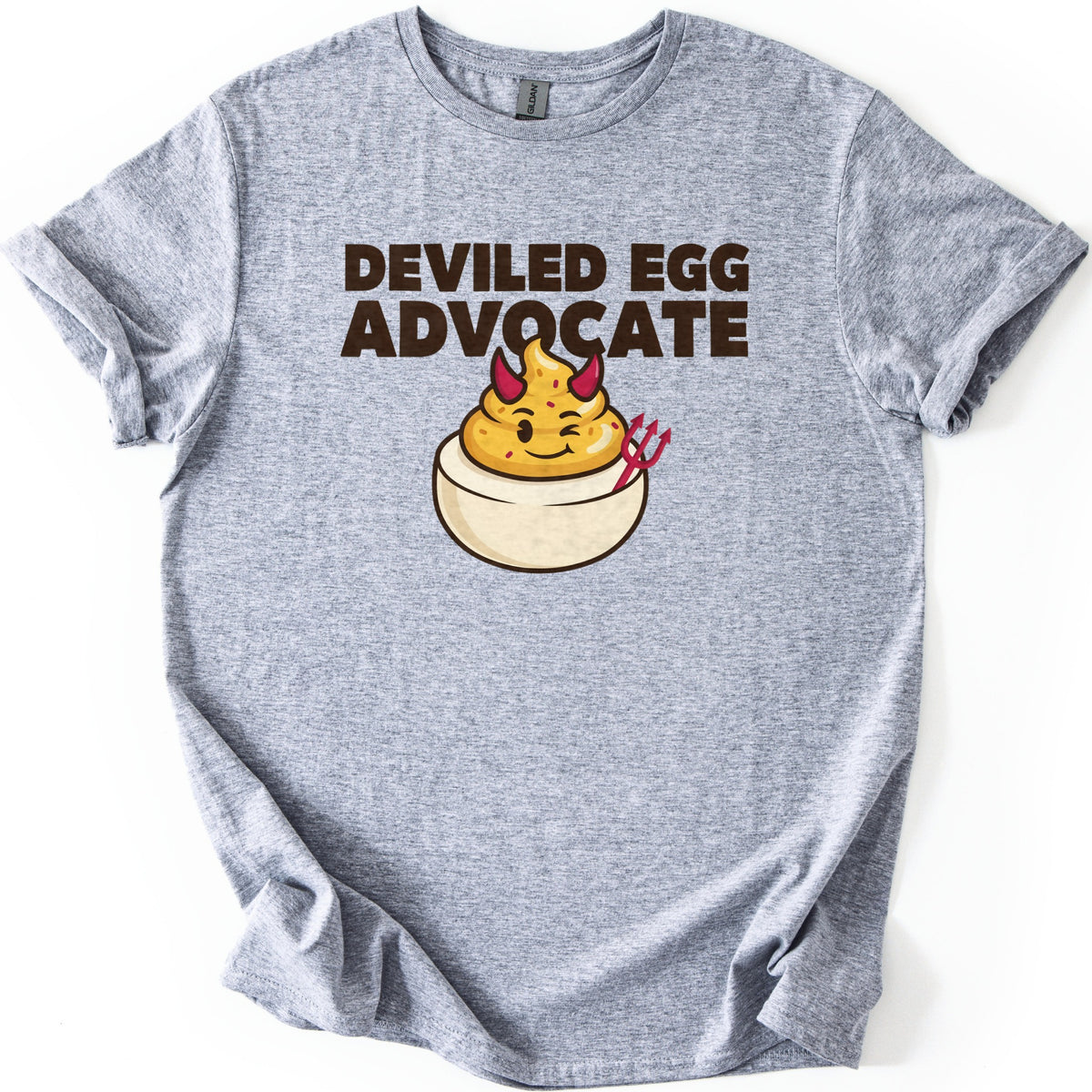 Deviled Egg Advocate Comfort Colors T-shirt in Sport Grey, laid flat. Bold dark brown text reads Deviled Egg Advocate above a cartoon deviled egg with red devil horns, a wink, and a small red pitchfork. The shirt has cuffed sleeves and lies on a white background.