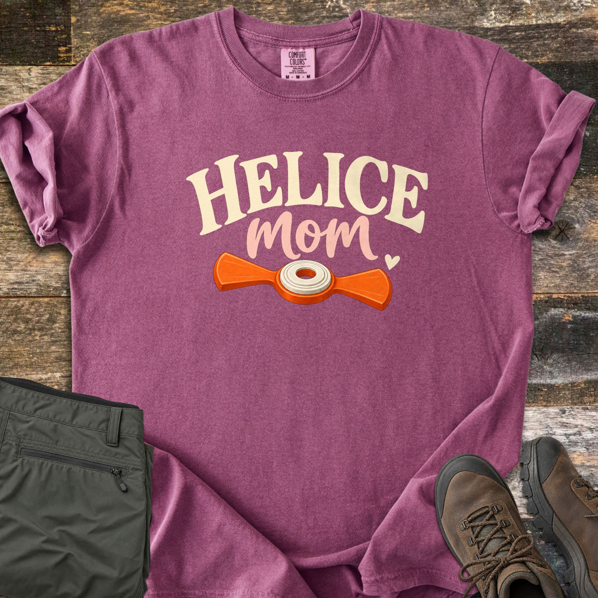 Helice Mom Comfort Colors T-shirt in Berry, laid flat. Cream letters spell HELICE over mom in pink script and a small heart, above an orange helice target with a white center ring. Olive pants and brown hiking boots sit beside it on rustic wood planks.