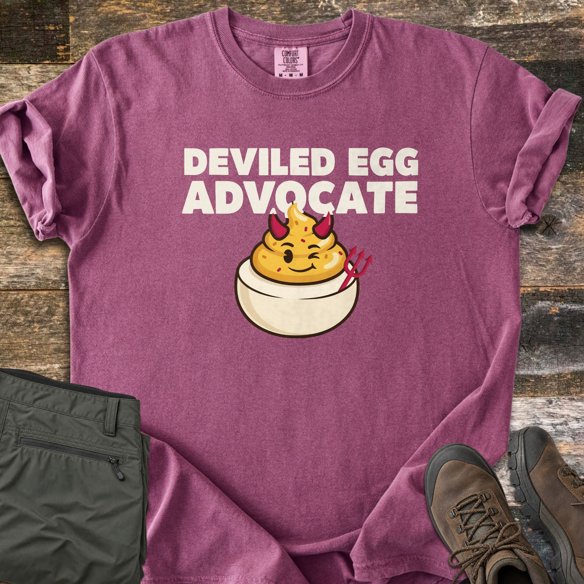 Deviled Egg Advocate Comfort Colors T-shirt in Berry, laid flat. Cream letters read Deviled Egg Advocate above a winking cartoon deviled egg with a piped yellow yolk, red devil horns and a red pitchfork. Olive hiking pants and brown boots lie beside it on rustic wood.