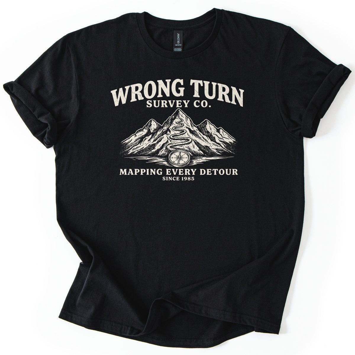 Wrong Turn Survey Co Comfort Colors T-shirt in Black, laid flat. Cream print reads Wrong Turn Survey Co. above sketched mountains with a winding trail up the tallest peak and a compass at its base, then Mapping Every Detour, Since 1985. Sleeves cuffed on a white background.