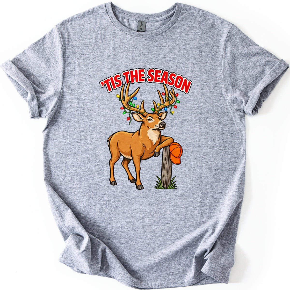 Buck Tis The Season Comfort Colors T-shirt in Sport Grey, laid flat. Red arched text reads 'TIS THE SEASON above a cartoon buck with colored Christmas lights strung on its antlers, leaning on a wooden post that holds an orange hunting cap. Sleeves rolled, on a white surface.