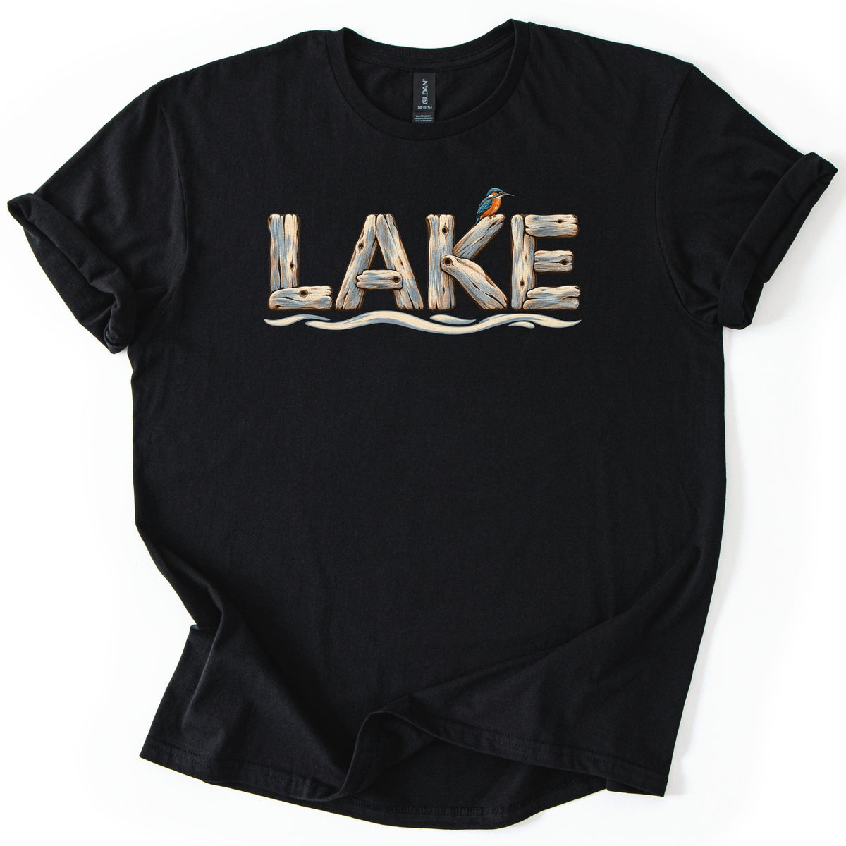 Lake Comfort Colors T-shirt in Black, laid flat. The word LAKE is spelled in weathered driftwood planks, with a small blue and orange kingfisher perched on the K and wavy water lines below. The shirt lies on white with rolled sleeves.
