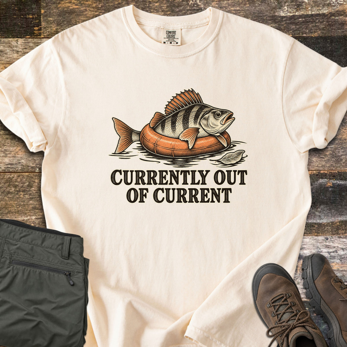 Currently Out of Current Comfort Colors T-shirt in Ivory, laid flat. A striped perch lounges on an orange inner tube floating in water beside a leaf, above the words Currently Out of Current. Olive hiking pants and brown hiking boots sit on weathered wood planks.