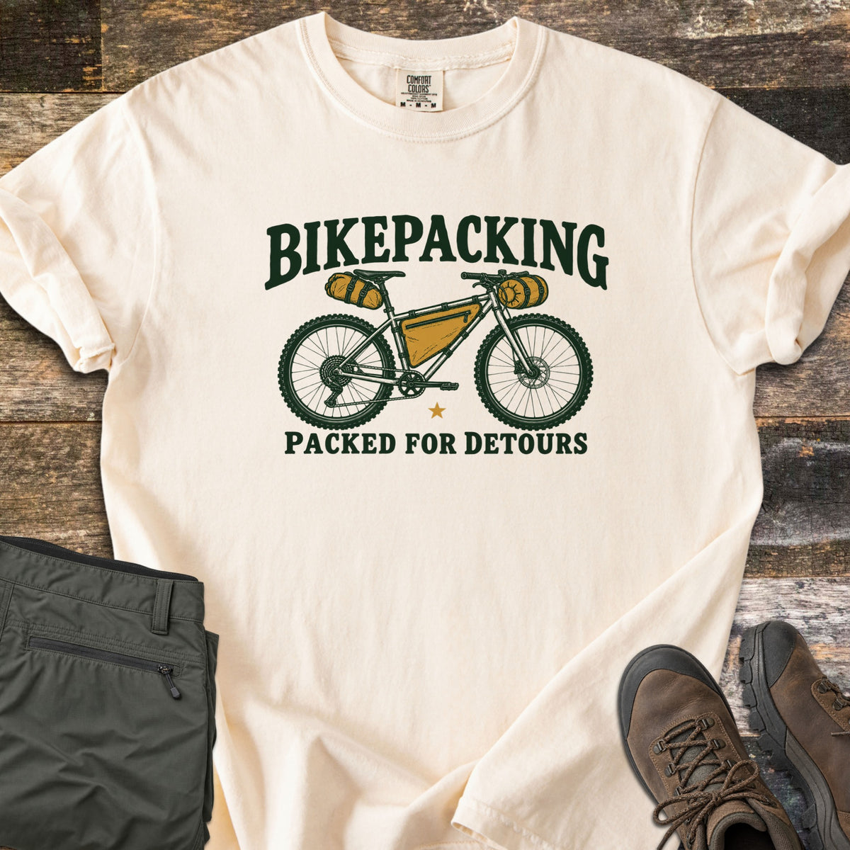Bikepacking Packed for Detours Comfort Colors T-shirt in Ivory, laid flat. A loaded bikepacking bike with yellow bags sits between the words Bikepacking and Packed for Detours in dark green. Folded olive pants and brown hiking boots lie beside it on weathered wood planks.