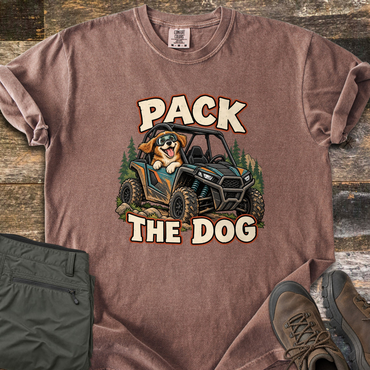 Pack the Dog Comfort Colors T-shirt in Espresso, laid flat. A cartoon dog in goggles rides in a muddy off-road side-by-side among pine trees, between the words Pack The Dog. Sleeves rolled, on weathered wood beside olive hiking pants and brown hiking boots.