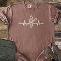 Kayak Heartbeat Comfort Colors T-shirt in Espresso, laid flat. A cream heartbeat line runs across the chest with a top-down kayak and paddle at its center. The shirt has rolled sleeves on rustic wood planks next to folded olive hiking pants and a pair of brown leather boots.