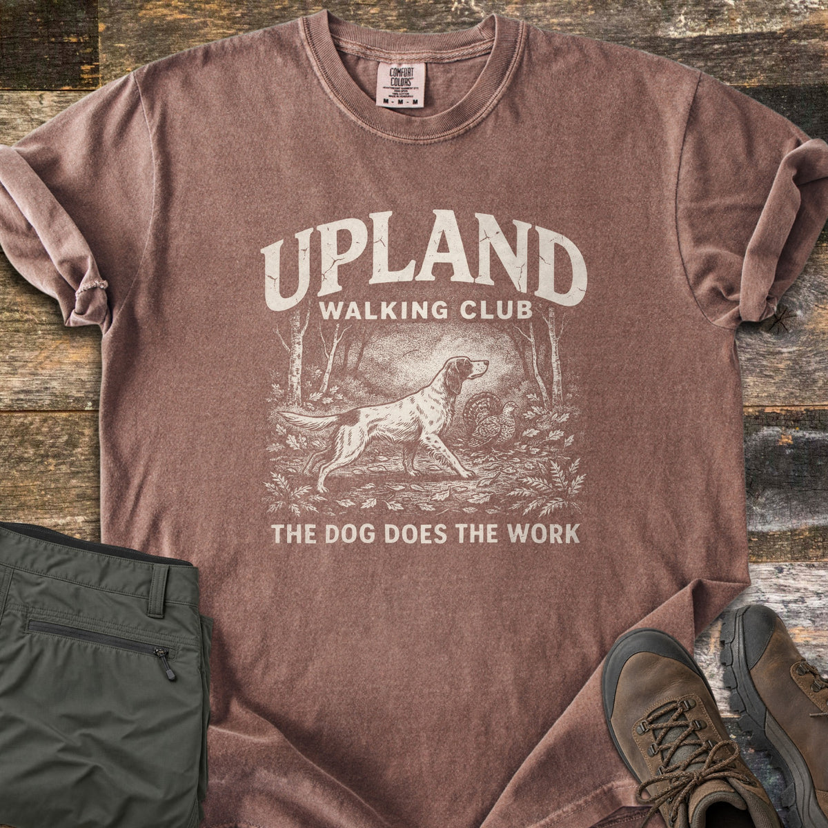 Upland Walking Club Comfort Colors T-shirt in Espresso, laid flat. A vintage-style sketch of a spaniel pointing at a grouse in the woods, under the words Upland Walking Club and above The Dog Does The Work. Olive pants and brown hiking boots sit beside it on weathered wood.