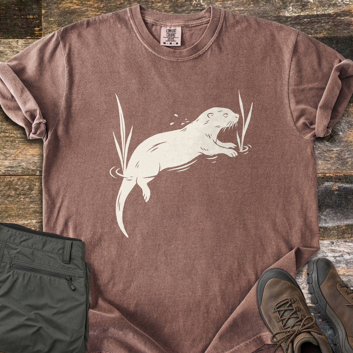 Swimming Otter Comfort Colors T-shirt in Espresso, laid flat. A cream print of an otter swimming between tall reeds, with ripples and splashing droplets. Sleeves are rolled; olive hiking pants and brown hiking boots sit on weathered wood planks.