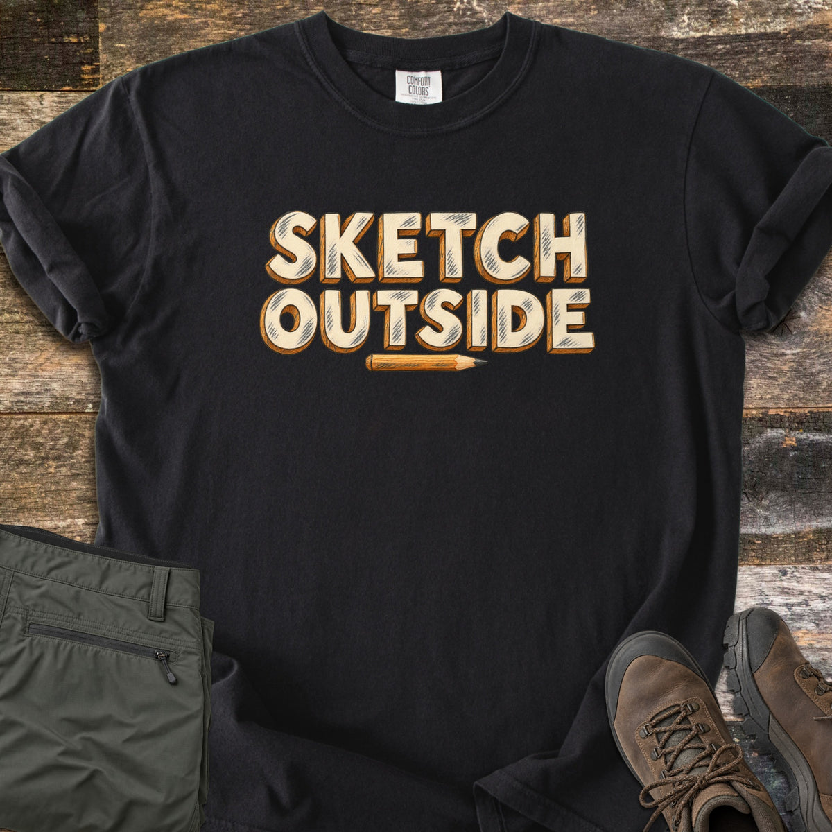 Sketch Outside Comfort Colors T-shirt in Black, laid flat. The words Sketch Outside in cream block letters with pencil-style shading and orange wood edges sit above a small yellow pencil. Olive hiking pants and brown hiking boots lie beside it on weathered wood planks.