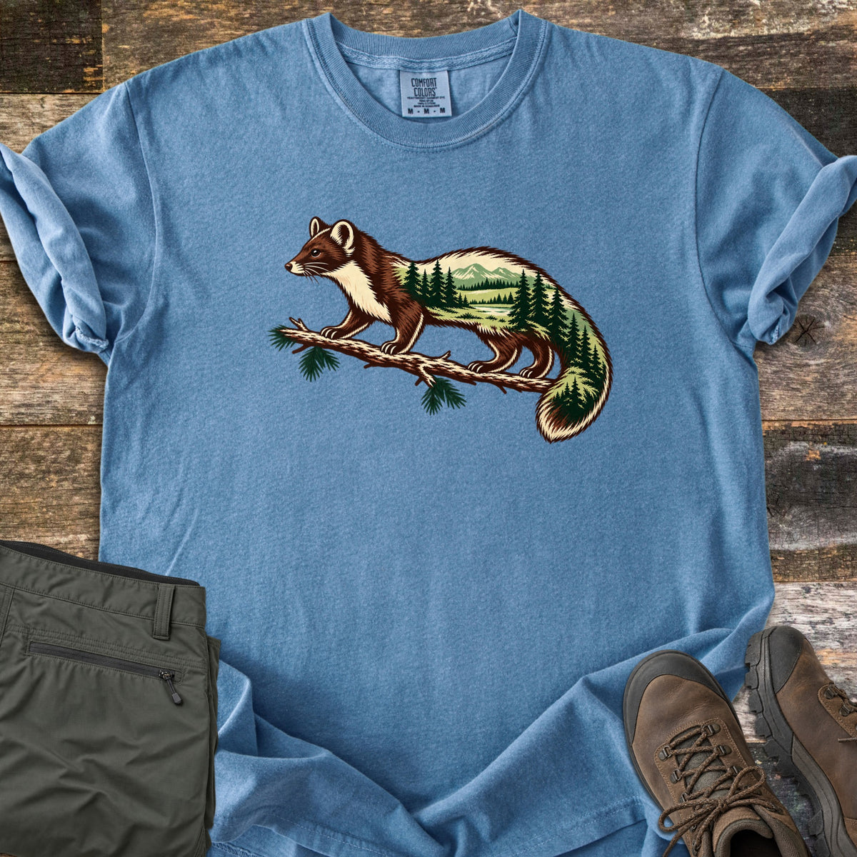 Pine Marten Forest Comfort Colors T-shirt in Blue Jean, laid flat. A pine marten stands on a pine branch, its body filled with a scene of evergreen trees, a lake and mountains. No words are printed. Shown on weathered wood with folded olive pants and brown hiking boots.