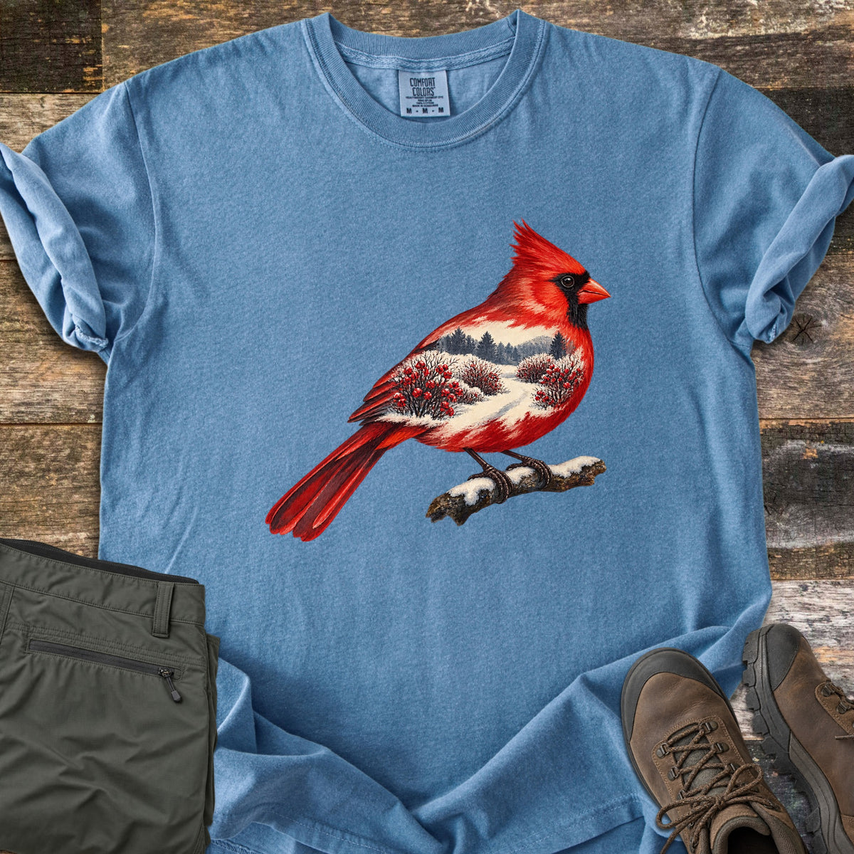 Winter Cardinal Comfort Colors T-shirt in Blue Jean, laid flat. A red cardinal perched on a snowy branch, its body filled with a snowy landscape of pine trees and red berry bushes. Rolled sleeves on weathered wood, with olive pants and brown hiking boots beside it.