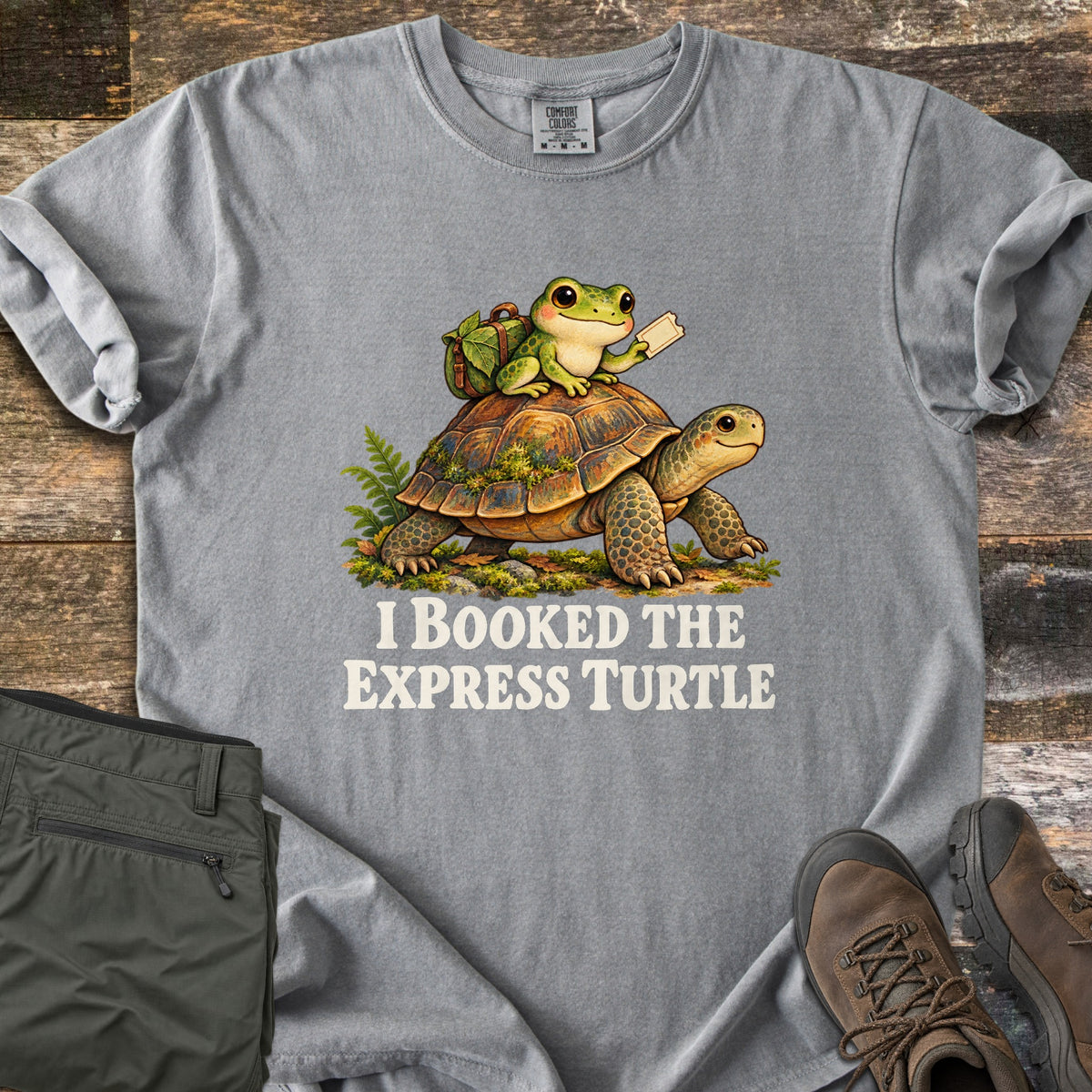 I Booked the Express Turtle Comfort Colors T-shirt in Grey, laid flat. A cartoon frog with a leafy backpack holds a ticket while riding a mossy tortoise, above the words I Booked the Express Turtle in cream. Shown on weathered wood with folded olive pants and brown hiking boots.