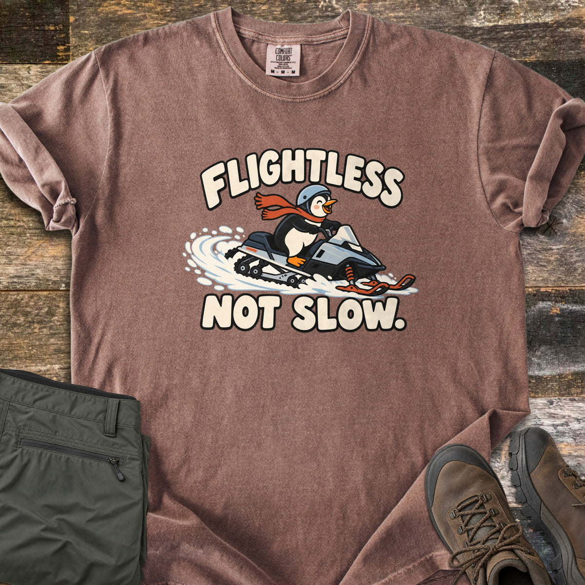 Flightless Not Slow Comfort Colors T-shirt in Espresso, laid flat. A cartoon penguin in a helmet and red scarf rides a snowmobile through snow between the words Flightless Not Slow. Folded olive pants and brown hiking boots sit beside it on weathered wood.