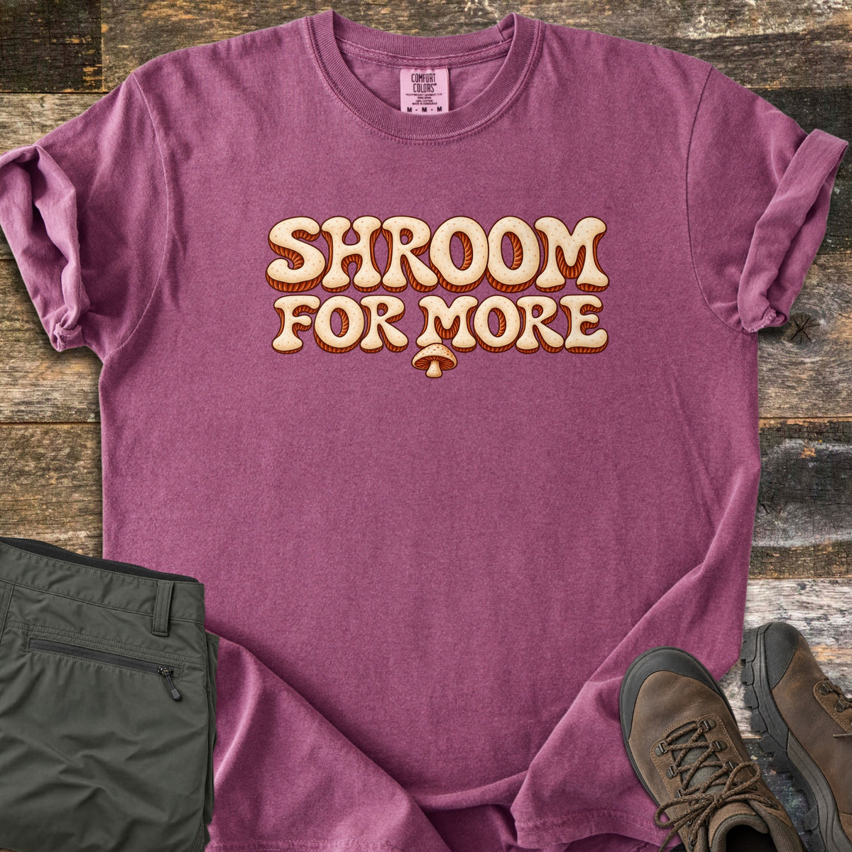 Shroom for More Comfort Colors T-shirt in Berry, laid flat. Retro cream bubble letters with rust-orange striped shadows read Shroom For More, with a small mushroom below. Sleeves are rolled on a rustic wood floor beside olive cargo pants and brown hiking boots.