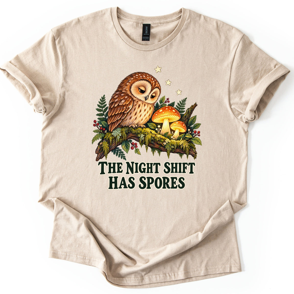 The Night Shift Has Spores Comfort Colors T-shirt in Sand, laid flat. A sleepy brown owl perches on a mossy branch beside two glowing orange mushrooms, with ferns, red berries and small stars, above the words The Night Shift Has Spores in dark green. Shown on white.