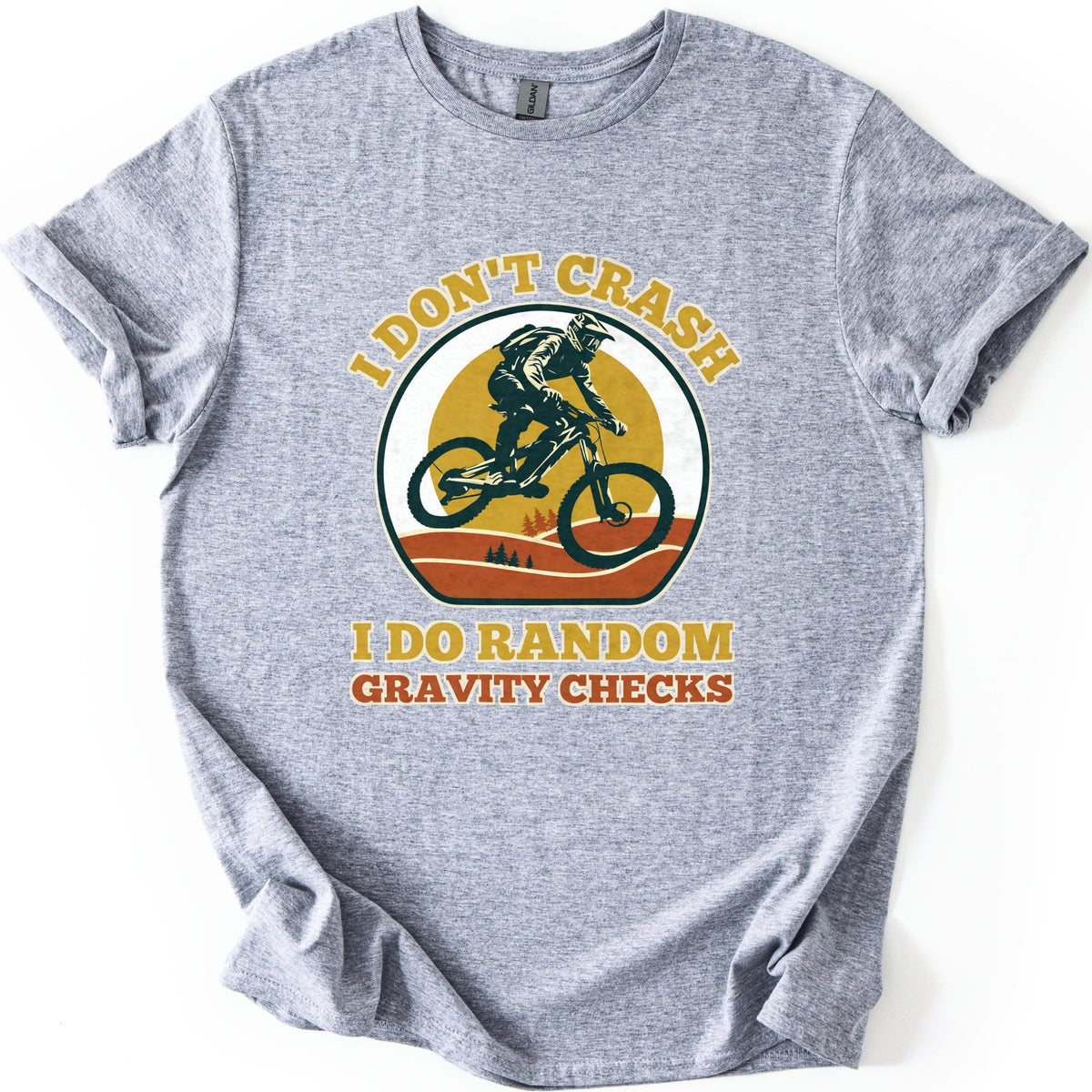 Random Gravity Checks MTB Comfort Colors T-shirt in Sport Grey, laid flat. A mountain biker in a full-face helmet jumps against a mustard sun over orange hills with pine trees, framed by the words I Don't Crash above and I Do Random Gravity Checks below. Sleeves rolled, on white.