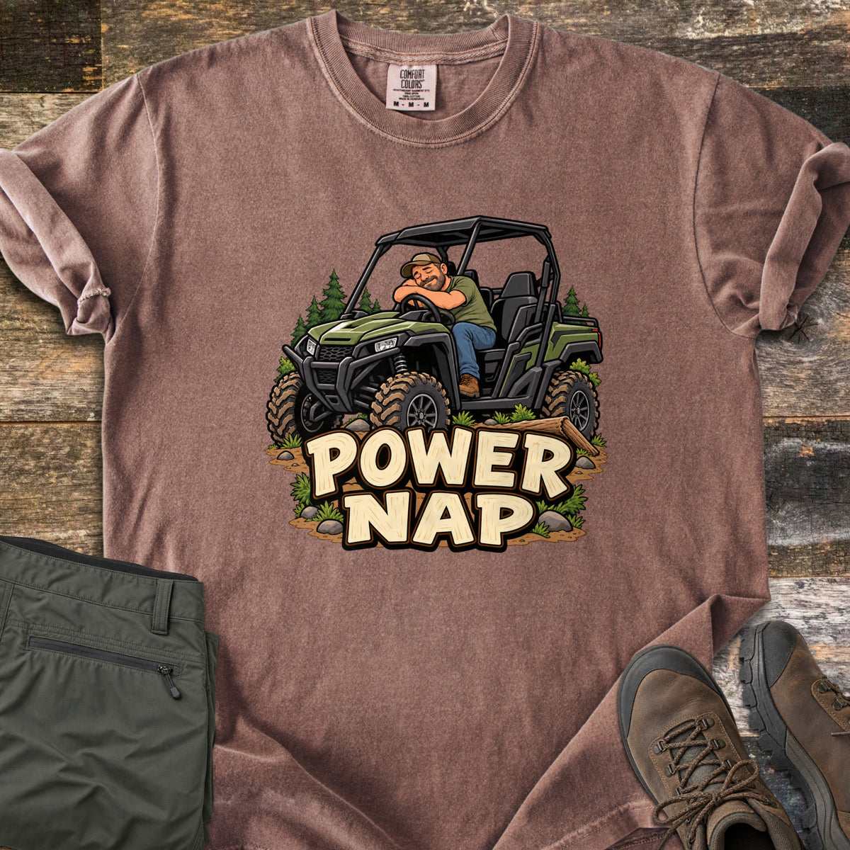 Power Nap Comfort Colors T-shirt in Espresso, laid flat. A cartoon man in a cap naps with his head on the steering wheel of a green side-by-side UTV among pine trees, above the words Power Nap. Folded olive pants and brown hiking boots sit on a weathered wood surface.