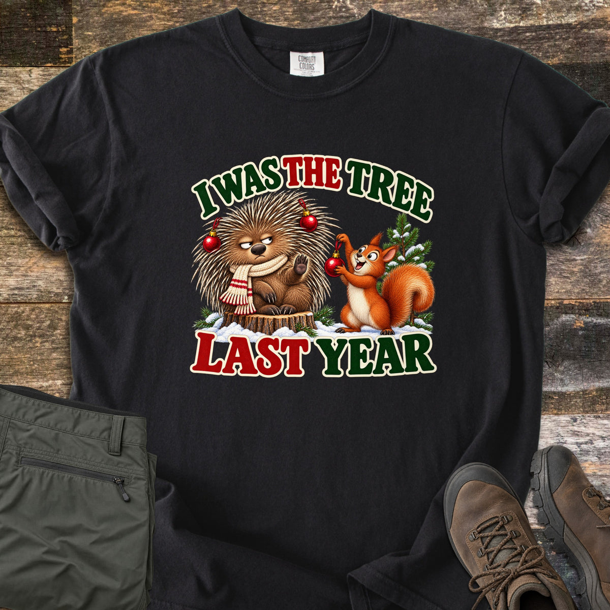I Was The Tree Last Year Comfort Colors T-shirt in Black, laid flat. A grumpy porcupine in a striped scarf sits on a stump with red ornaments in its quills as a smiling squirrel hangs another, between the words I Was The Tree Last Year. Olive pants and brown boots lie on wood planks.