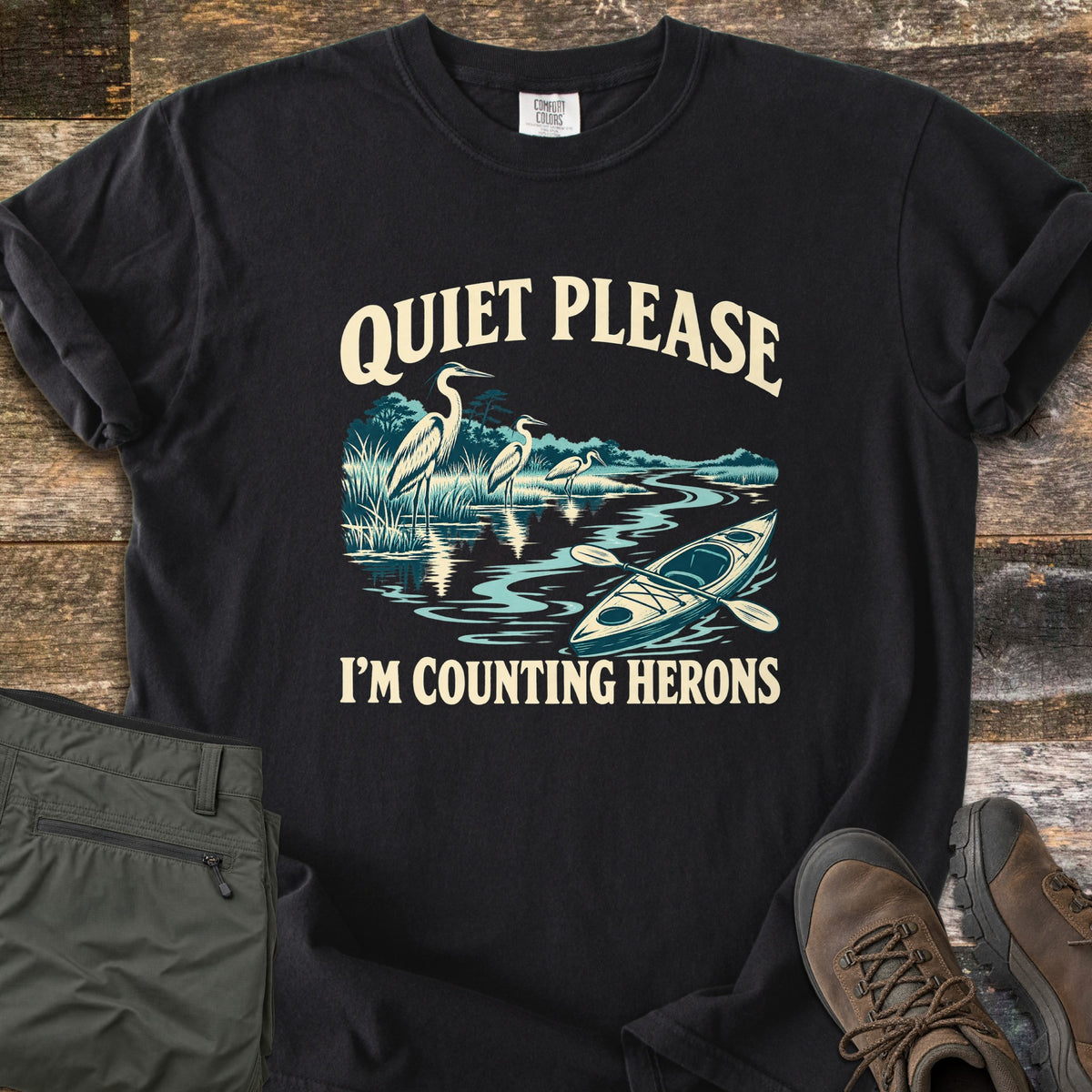 Counting Herons Comfort Colors T-shirt in Black, laid flat. Printed text Quiet Please arches over three herons standing in marsh grass by a winding river with a kayak and paddle, above I'm Counting Herons. Olive cargo pants and brown hiking boots sit on weathered wood.