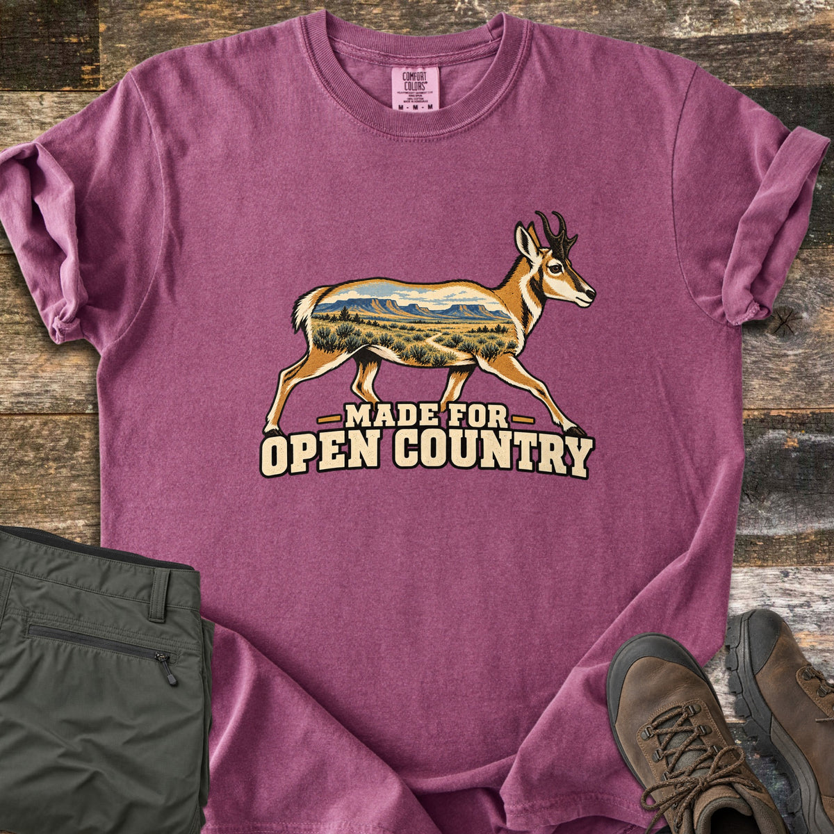 Made for Open Country Comfort Colors T-shirt in Berry, laid flat. A running pronghorn whose body is filled with a desert scene of mesas, sagebrush and a trail, above the words Made for Open Country. Sleeves are rolled on a wood background with green pants and brown hiking boots.