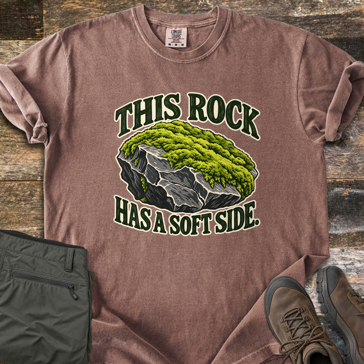 This Rock Has a Soft Side Comfort Colors T-shirt in Espresso, laid flat. An illustrated gray boulder topped with thick green moss sits between the words This Rock above and Has a Soft Side. below. Olive hiking pants and brown hiking boots lie beside it on weathered wood planks.