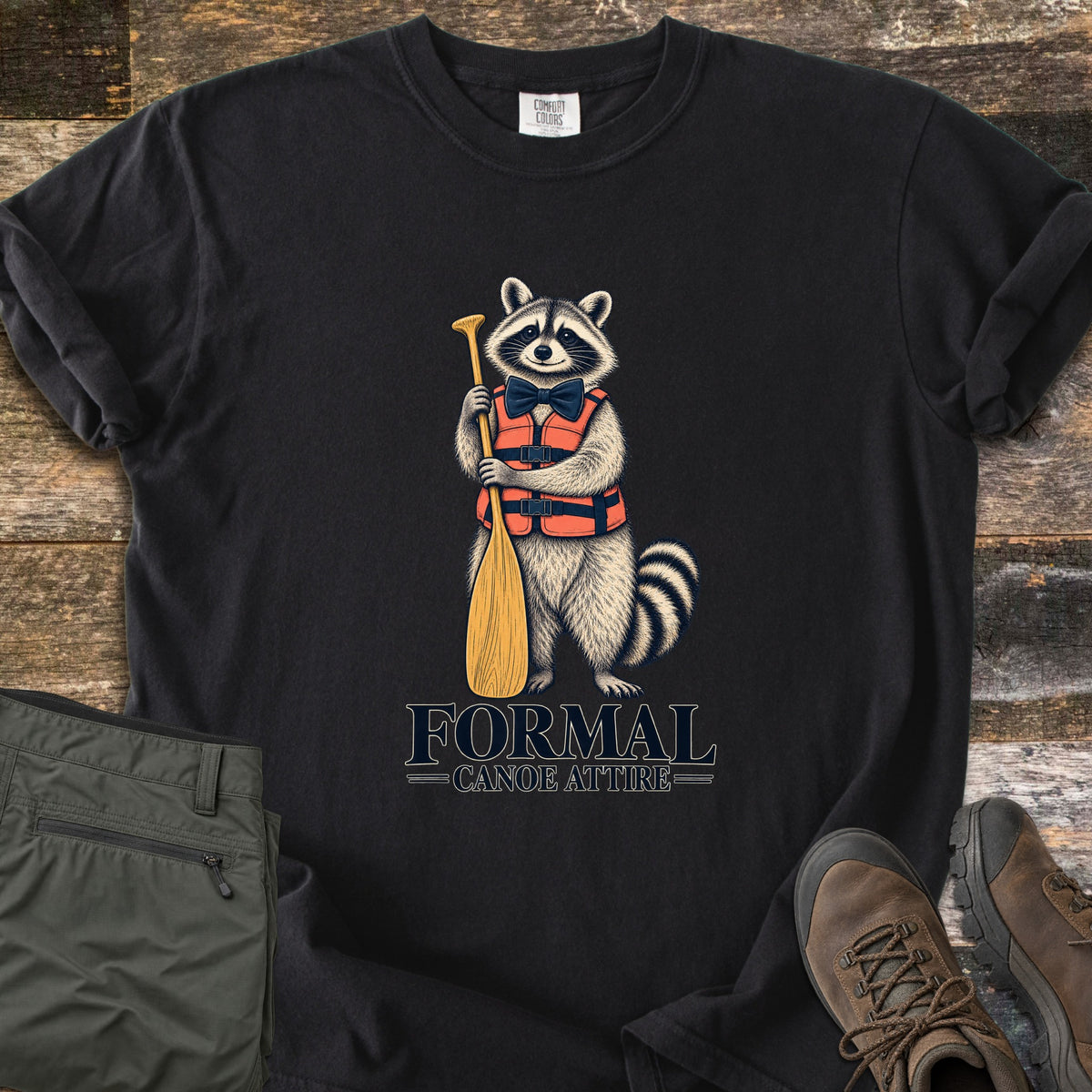 Formal Canoe Attire Comfort Colors T-shirt in Black, laid flat. A raccoon in an orange life vest and navy bow tie stands holding a wooden canoe paddle above the words Formal Canoe Attire. Olive hiking pants and brown hiking boots sit beside it on weathered wood planks.