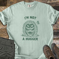 I'm Not A Hugger Comfort Colors T-shirt in Bay, laid flat. A green sketch of a grumpy hedgehog with crossed arms and a butterfly beside it, between the words I'M NOT A HUGGER. Shown on wood boards with folded olive hiking pants and brown hiking boots.