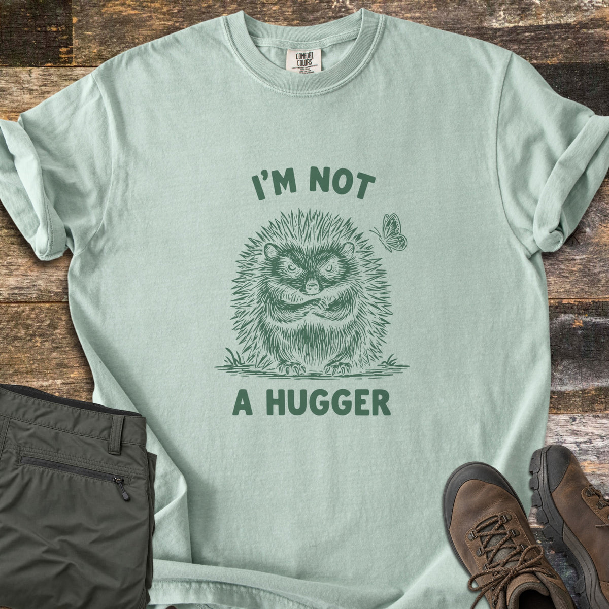 I'm Not A Hugger Comfort Colors T-shirt in Bay, laid flat. A green sketch of a grumpy hedgehog with crossed arms and a butterfly beside it, between the words I'M NOT A HUGGER. Shown on wood boards with folded olive hiking pants and brown hiking boots.