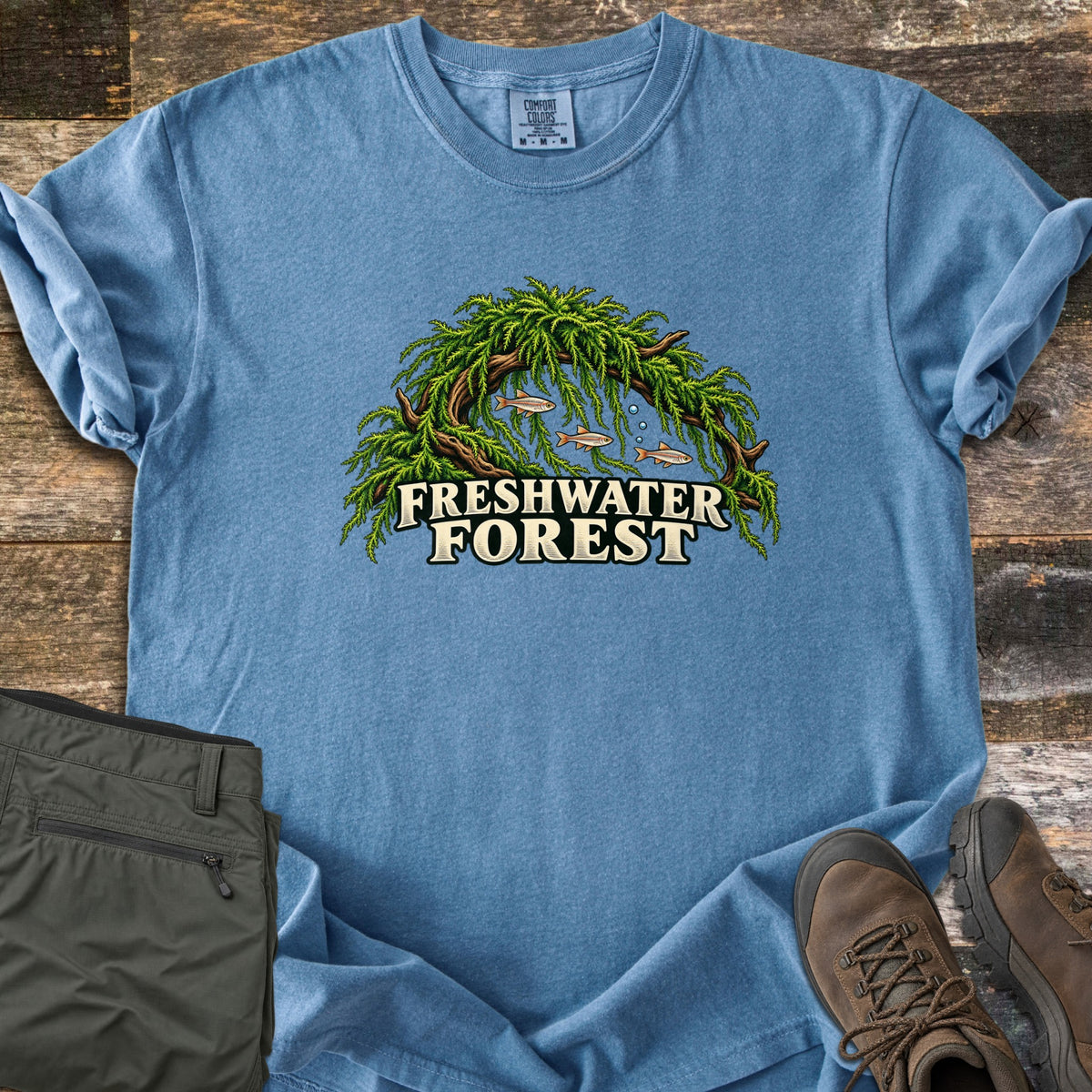 Freshwater Forest Comfort Colors T-shirt in Blue Jean, laid flat. Green moss hangs from a curved driftwood branch over three small fish and rising bubbles, above the words Freshwater Forest. Folded olive pants and brown hiking boots sit on rustic wood boards.