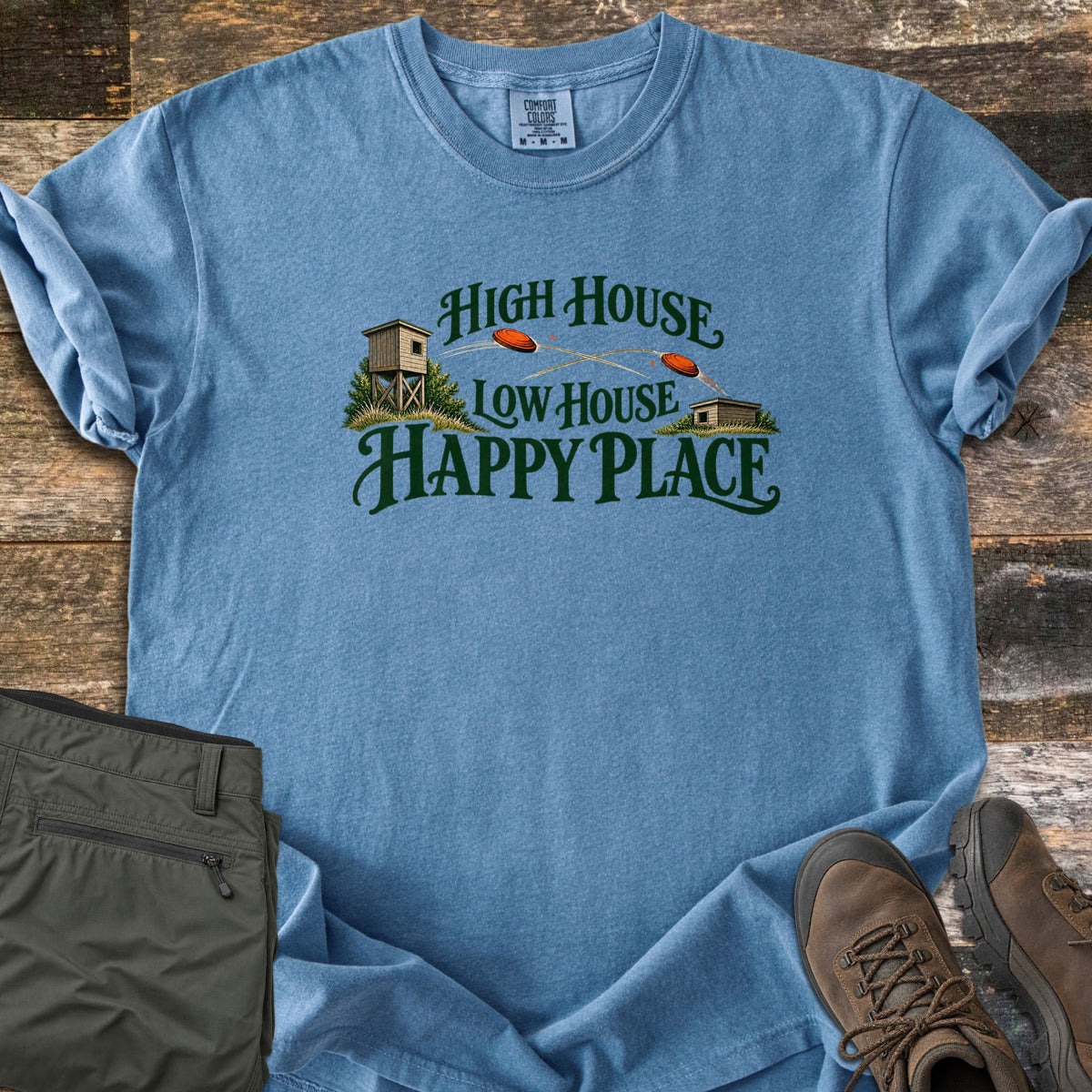 High House Low House Happy Place Comfort Colors T-shirt in Blue Jean, laid flat. Green lettering reads High House Low House Happy Place around two skeet houses with orange clay targets flying between them. Olive cargo pants and brown hiking boots lie beside it on weathered wood planks.