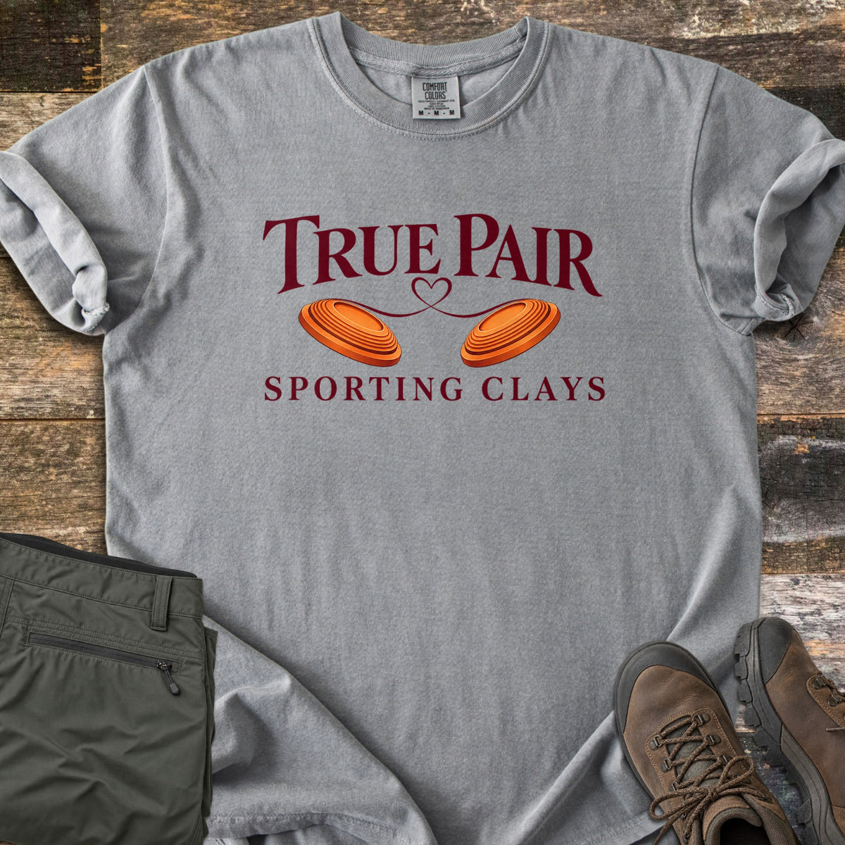 True Pair Comfort Colors T-shirt in Grey, laid flat. Dark red text reads True Pair above two orange clay targets joined by a looping heart line, with Sporting Clays below. Sleeves are rolled; olive pants and brown hiking boots sit on weathered wood.