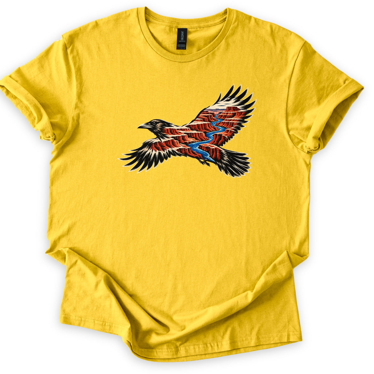 Red Canyon Raven Comfort Colors T-shirt in Daisy, laid flat. A black raven in flight, its body and wings filled with red canyon cliffs and a winding blue river. No words are printed. The shirt lies on a white surface with rolled sleeves and a loosely folded hem.