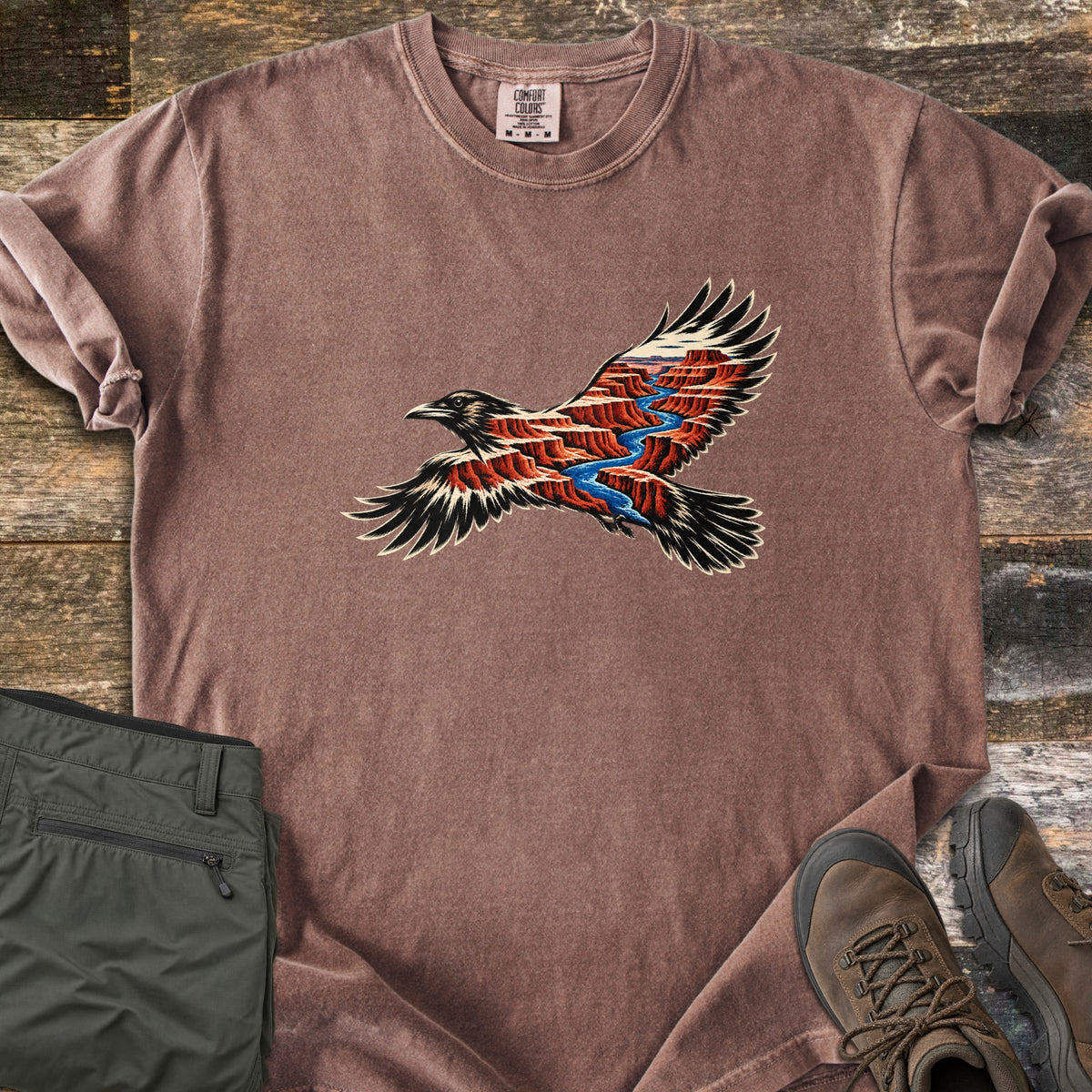 Red Canyon Raven Comfort Colors T-shirt in Espresso, laid flat. A flying black raven whose body and wings hold a scene of red canyon walls and a winding blue river. Sleeves rolled, on dark wood planks beside folded olive pants and brown hiking boots.