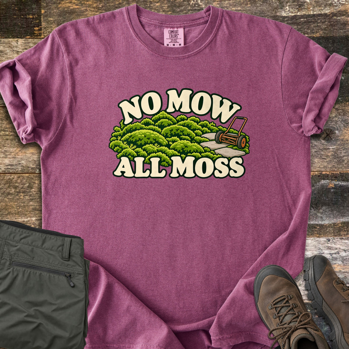 No Mow All Moss Comfort Colors T-shirt in Berry, laid flat. Cream letters read No Mow above and All Moss below a scene of green moss mounds, flat stepping stones and an orange reel mower. Rolled sleeves rest on dark wood planks next to olive pants and brown boots.