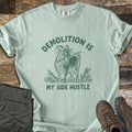 Demolition Is My Side Hustle Comfort Colors T-shirt in Bay, laid flat. Green print of a shaggy goat holding a broken board in its mouth among splintered wood, with the words Demolition Is arched above and My Side Hustle below. Olive pants and brown hiking boots lie on weathered wood.